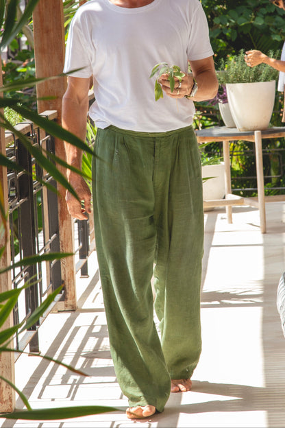 Person wearing a white t-shirt and green pants standing outdoors with plants and furniture in the background.
